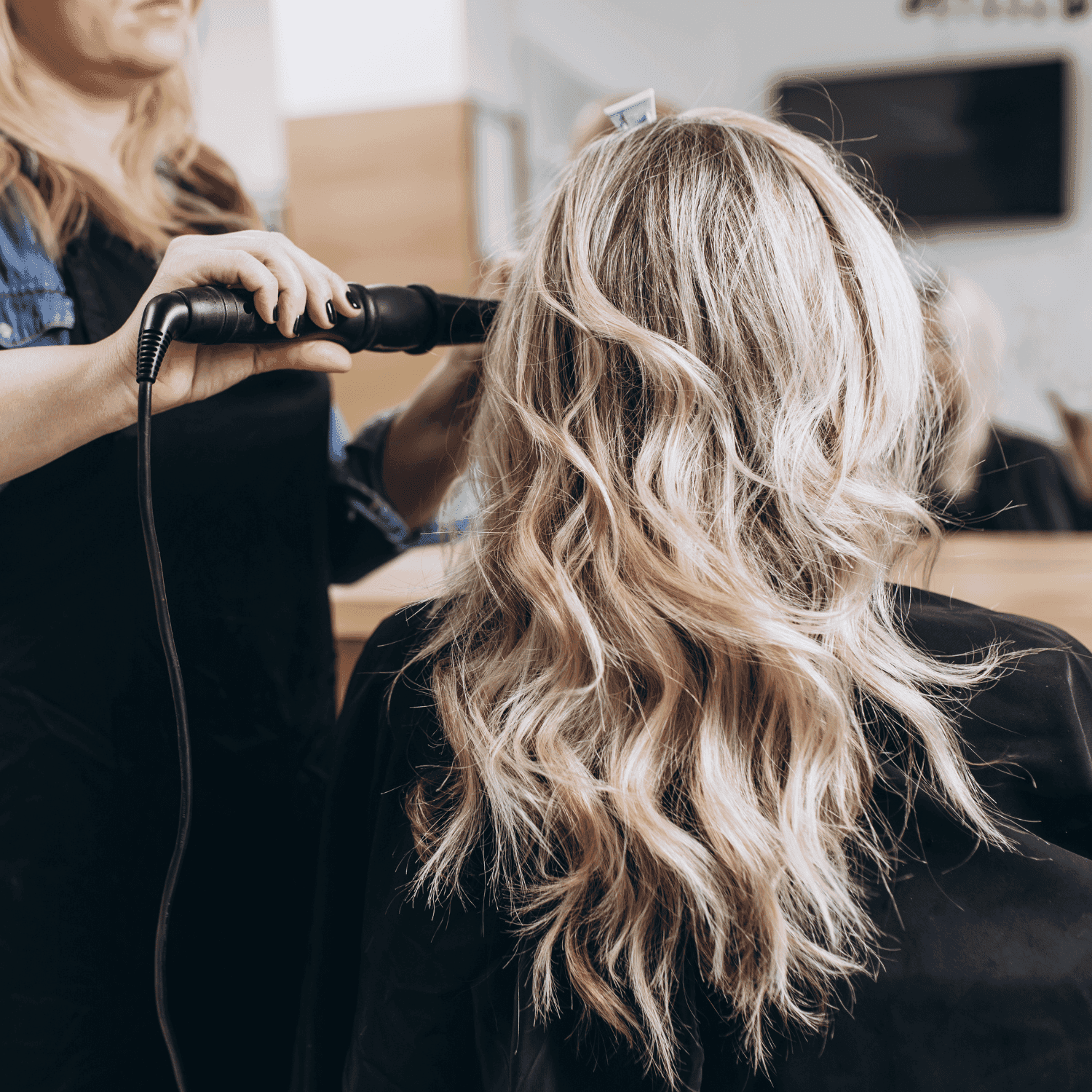 Hairstylist curling a client's long, wavy blonde hair in a salon setting.